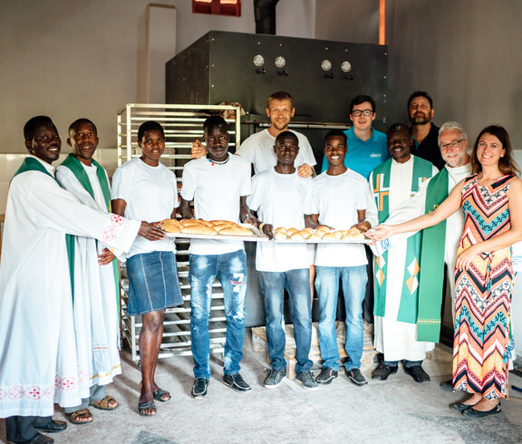 Die Bäcker der neuen Bäckerei in Uganda stehen gemeinsam mit Förder*innen dem jetzigen Bischof von Kärnten Josef Marketz, sowie Bereichsleiterin der Caritas Kärnten für die Auslandshilfe Alexandra Blattnig Rull vor den großen Backöfen und einem Blech voll frisch gebackenen Brot.