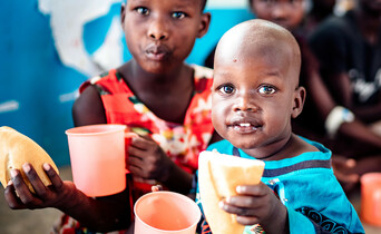 Zwei Kinder in Uganda sitzen in der Schule und haben ein frischgebackenes Brot sowie bunte Trinkbecher in der Hand.