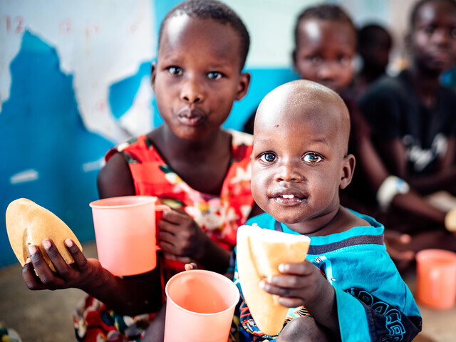 Zwei Kinder in Uganda sitzen in der Schule und haben ein frischgebackenes Brot sowie bunte Trinkbecher in der Hand.