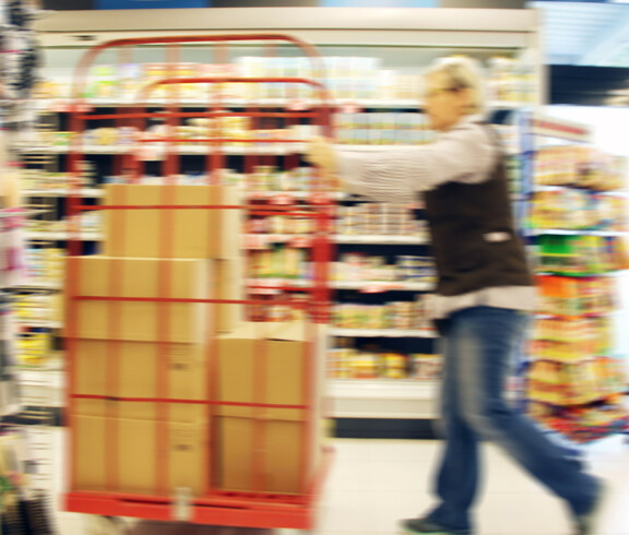 Eine SPAR Supermarkt Perspektive Handel-Mitarbeiterin fährt mit einem Rollwagen und neuen Waren durch den Gang des Supermarktes.