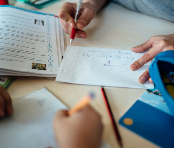 Man sieht die Hände eines Lernhelfers, der einem Schüler gerade eine Aufgabe Schritt für Schritt anhand einer Rechnung auf einem Blatt erklärt.