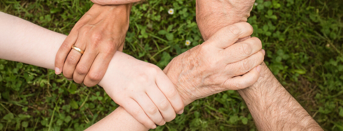 Sechs Hände nehmen sich gegenseitig am Handgelenk und formen so über einer grünen Wiese einen Kreis.