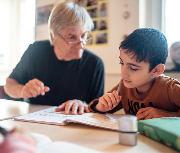 Ein Mann sitzt mit einem Jungen am Tisch auf dem auch Heft, Spitzer und Federpenal liegen. Er erklärt dem Jungen gerade eine Aufgabe. Der Junge sieht konzentriert in das Heft.