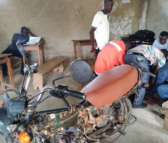 Ein altes Motorrad steht in der Mitte, im Hintergrund arbeiten junge Männer am Motor um diesen wieder zu reparieren.