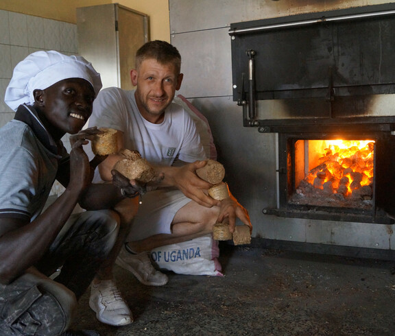 Ein Mitarbeiter der Bäckerei und der österreichisches Bäcker Josef knien vor dem großen Backofen und halten das Heizmaterial mit der Ofen beheizt wird in den Händen.