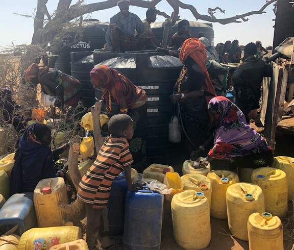 Menschen und Kinder stehen mit vielen gelben Kanistern vor dem großen schwarzen Wassertank.