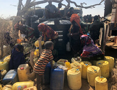 Menschen und Kinder stehen mit vielen gelben Kanistern vor dem großen schwarzen Wassertank.