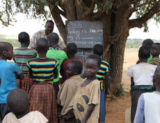 An einem Baum ist eine Tafel angebracht, die afrikanischen Kinder stehen vor diesem und haben mit einem Lehrer Unterricht.