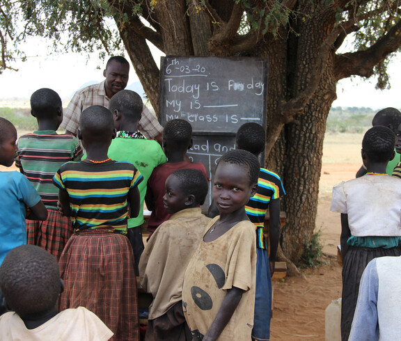 An einem Baum ist eine Tafel angebracht, die afrikanischen Kinder stehen vor diesem und haben mit einem Lehrer Unterricht.