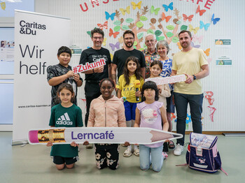 Kinder, Freiwillige und Mitarbeiter der Lerncafés feiern gemeinsam mit Caritasdirektor Ernst Sandriesser und Antenne Kärnten Mitarbeiter die erfolgreiche Antenne-Schulstartaktion. Gruppe aus Kindern und Erwachsenen im Lerncafé mit Schildern zu #Zukunft und #Chancen bei der Antenne-Schulstartaktion.