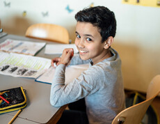 Ein Schüler sitzt an mit seinem Federpenal, Heft und einem Übungszettel am Tisch und lächelt in die Kamera.