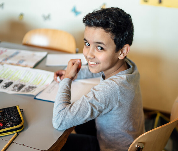 Ein Schüler sitzt an mit seinem Federpenal, Heft und einem Übungszettel am Tisch und lächelt in die Kamera.