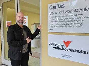 Hereinspaziert: Schulleiter Wolfgang Kogler lädt interessierte Pflichtschulabgänger*innen zum Besuch der Fachschule für Sozialberufe mit Pflegevorbereitung ein. Schulleiter steht vor Eingang der Caritas-Schule für Sozialberufe in Wolfsberg und zeigt auf das Schulschild.