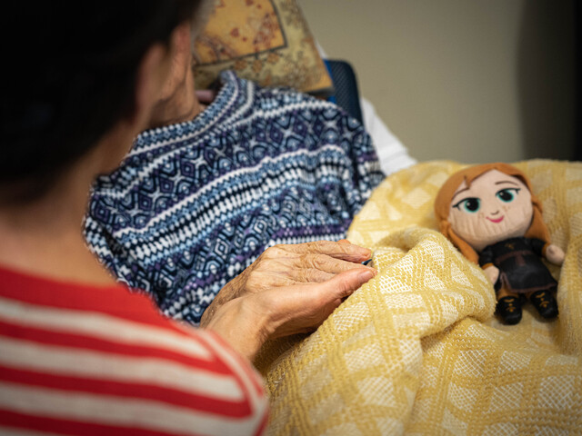 Unsere mobilen Hospizbegleiter*innen stehen sterbenden Menschen und ihren Angehörigen zur Seite. Kostenlos, dank vieler Spenden.  Eine ältere Person im Pflegebett wird von einer Angehörigen gehalten, eine Stoffpuppe liegt auf der gelben Decke.