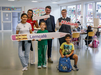 Unterstützen Kinder auf ihrer Bildungsreise: Dr. Heinz Paar, Caritasdirektor Ernst Sandriesser und Magistra Roberta Striedinger (Caritas) mit Schüler*innen. Zwei Männer, eine Caritas-Mitarbeiterin und drei Kinder halten Lerncafé-Schilder in einem Lernraum.