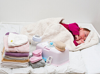 Ein Baby liegt eingewickelt in eine Babydecke auf einem Wickeltisch, daneben liegt die Erstausstattung für das Baby.