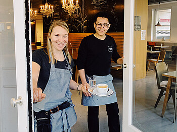 Die Servicechefin und ein Kellner des magdas LOKAL am Stauderplatz in Klagenfurt halten eine Tasse Kaffee in der Hand und halten die Eingangstür offen.