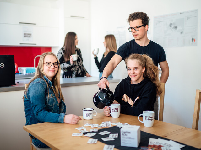 Zwei junge Frauen sitzen zu einem Coffee to help am Esstisch und ein junger Mann schenkt ihnen Kaffee ein. Im Hintergrund unterhalten sich zwei weitere Mädchen mit einer Tasse Kaffee in der Hand.