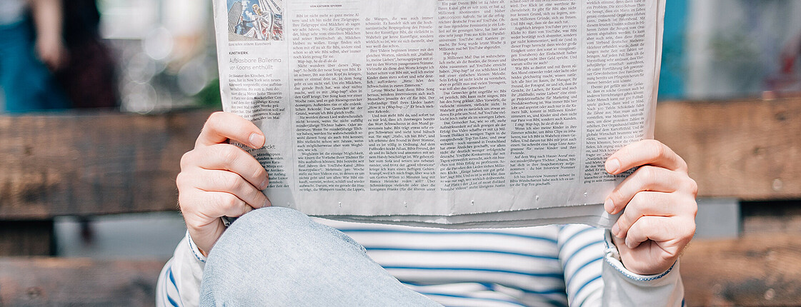 Ein Herr in blauem Streifen-Pullover und hellblauer Jeanshose hält eine Zeitung vor sein Gesicht.